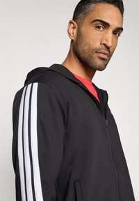 Čierna mikina na zips s bielymi vertikálnymi pruhmi na rukávoch, vyrobená z mäkkého materiálu. Má predné vrecko a kapucňu. Model nosí červené tričko pod mikinou.