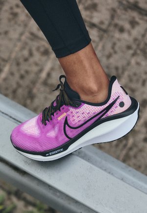 Stopa w różowo-fioletowym butach do biegania Nike ZoomX stąpa po metalowej szynie, osoba ubrana w czarne legginsy.