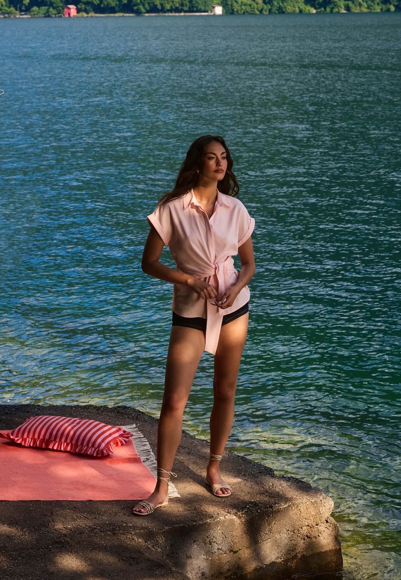Femme portant une chemise rose et un short noir, debout sur un bord rocheux au bord d’un lac avec un tapis corail et un coussin rayé à proximité.