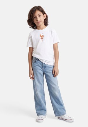 Barn med mellem-langt hår iført en hvid t-shirt med rødt logo, lyseblå jeans og hvide sneakers, stående mod en ensfarvet hvid baggrund.