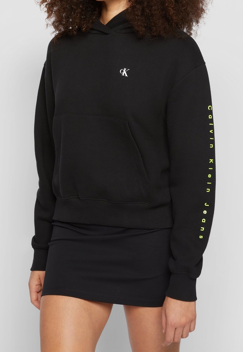 Haut de hoodie noir court avec le logo blanc "cK" sur la poitrine et le texte néon jaune "Calvin Klein Jeans" sur la manche, porté avec une mini jupe noire.