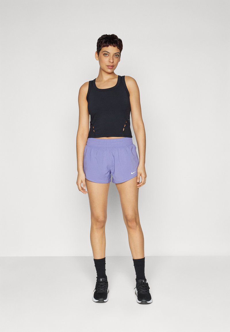 Schwarzes, kurzes Tanktop mit seitlichen Ausschnitten, kombiniert mit helllilafarbenen Sportshorts mit elastischem Bund und weißem Logo. Schwarze Turnschuhe.