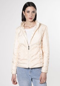 Giacca trapuntata color pesco chiaro con discreti pois bianchi, colletto alto, chiusura con zip e maniche aderenti. Indossata sopra una maglietta bianca.