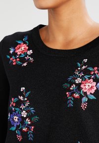 Pull noir avec broderies florales colorées en rose, bleu et vert, col rond et tissu texturé.