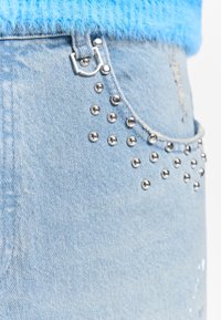 Poche de jean en denim bleu clair avec des clous en métal argenté et une petite déchirure près du bord, associée à un pull en maille duveteuse bleu vif.