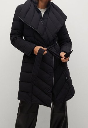 Personne portant un manteau matelassé noir longueur genou avec un large col, tenant la ceinture du manteau à la taille, et un pantalon noir dans un environnement neutre.