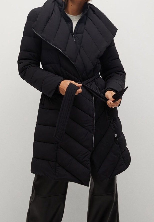 Personne portant un manteau matelassé noir longueur genou avec un large col, tenant la ceinture du manteau à la taille, et un pantalon noir dans un environnement neutre.