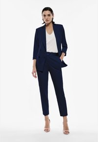 Completo su misura blu navy con giacca monopetto e pantaloni slim-fit, abbinato a un top bianco e décolleté nude con cinturini.
