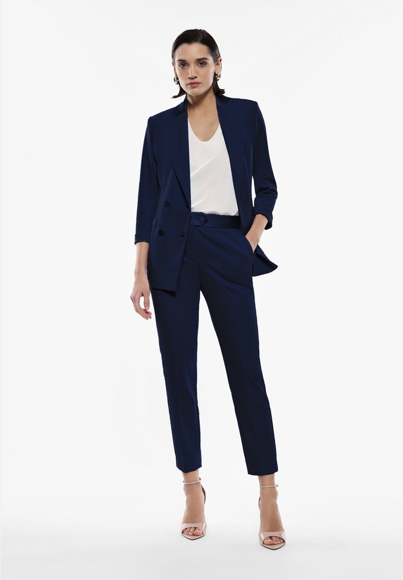 Completo su misura blu navy con giacca monopetto e pantaloni slim-fit, abbinato a un top bianco e décolleté nude con cinturini.