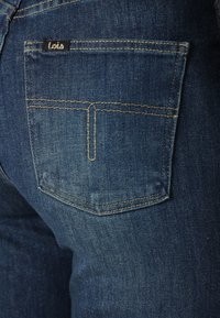 Sötétkék farmernadrág, hátul fekete logós címkével ellátott zsebbel és kontrasztos sárga varrással. Texturált anyag, enyhe kopással.