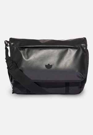 Sac messager noir avec bandoulière réglable, poche zippée avant et logo trèfle embossé sur le rabat.