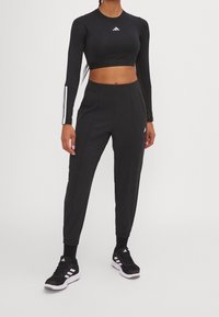 Zwarte lange mouwen crop top met een wit logo, gecombineerd met zwarte tapered broek met voorplooien en zwarte sport schoenen met witte strepen.
