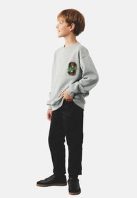 Szara bluza z kolorową haftowaną naszywką na piersi, w zestawieniu z czarnymi spodniami i czarnymi sneakersami, stojąca na białym tle.
