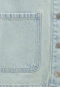 Närbild av en ljusblå denimjackas ficka med synliga sömmar och tre metallknappar på sidan.