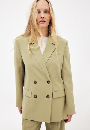 Blonde Frau in übergroßem hellolivgrünem, doppelt geknöpftem Blazer mit braunen Knöpfen über weißem Hemd, vor weißem Hintergrund stehend.