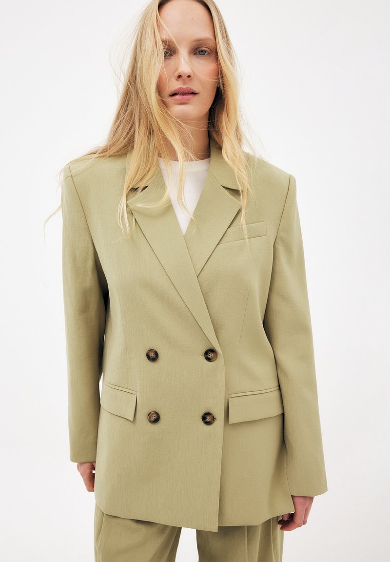 Vaalea nainen, joka käyttää ylisuurta vaaleanoliivinväristä kaksirivistä bleiseriä, jossa on ruskeat napit, valkoisen paidan päällä, seisoo valkoista taustaa vasten.