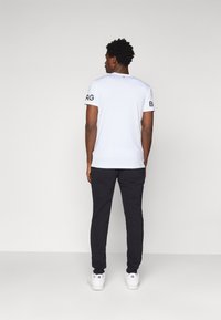 T-shirt branca de manga curta com letras pretas nas mangas, combinada com calças pretas e sapatilhas brancas, vista por trás.