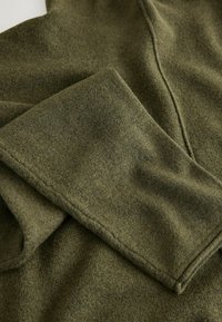 Zielony dzianinowy materiał o miękkiej teksturze, z złożonym rękawem i detalami szwów, pokazany w zbliżeniu.