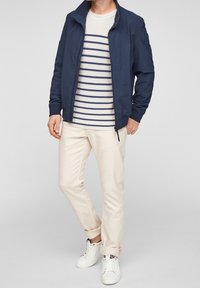 Veste marine à fermeture éclair, pull rayé crème et marine, pantalon beige clair et baskets blanches, avec revers de manches retroussés et design épuré.