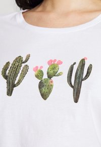 White T-shirt featuring cactus designs in green with pink flower accents, displaying three distinct shapes and textures.
