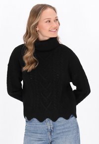 Schwarzer Strickpullover mit Rollkragen, der ein strukturiertes Zopfmuster und einen gewellten Saum aufweist. Kombiniert mit hellen Jeans.