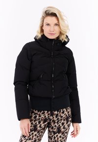 Mujer rubia con una chaqueta acolchada negra con bolsillos con cremallera y pantalones de leopardo estampados, de pie contra un fondo blanco.
