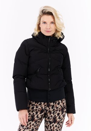 Blonde vrouw in een zwarte pufferjack met ritssluitingen en geprinte panterbroek, staand tegen een witte achtergrond.