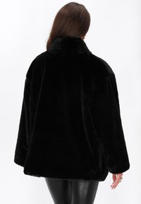 Donna che indossa una giacca nera in pelliccia sintetica e pantaloni in pelle neri, mostrata di spalle su uno sfondo neutro.