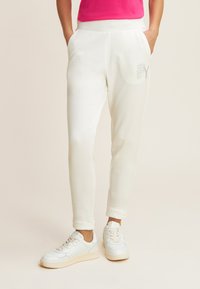 Pantalones de chándal blancos con un diseño ajustado, que cuentan con bolsillos laterales y un logo brillante de rhinestones en el muslo izquierdo. Acompañados de zapatillas blancas.
