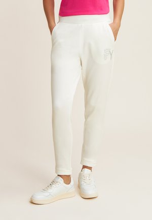 Pantaloni da tuta bianchi con un design affusolato, dotati di tasche laterali e un logo in strass lucido sulla coscia sinistra. Combinati con sneaker bianche.