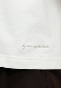 Λευκό ύφασμα με διακριτικό κεντημένο κείμενο "by marcopelo denim" κοντά στην εσοχή πάνω από μαύρο ύφασμα.