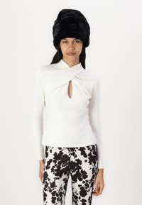 White long-sleeve top with a twisted neckline and a keyhole detail, paired with black and white spotted patterned pants and a black fur hat.