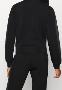 Person trägt schwarzen langärmligen Oberteil und Hose mit dezenten Logo-Streifen an Ärmeln und Seiten, steht mit dem Rücken zur Kamera.