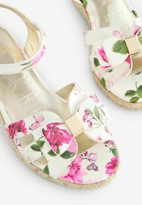 Płaskie sandały z kwiatowym wzorem, z paskiem na kostce, w kremowej bazie i akcentem w postaci kokardy. Złote detale i teksturowana podeszwa espadryli.