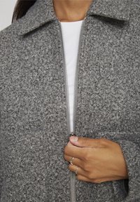 Szara, teksturowana kurtka na zamek błyskawiczny z kołnierzem, wykonana z materiału przypominającego wełnę, z gładkim zamkiem. Ręka umieszczona blisko zamka.