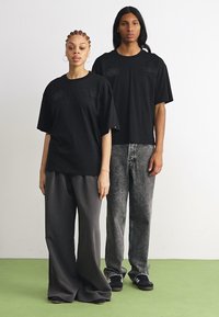 Tricouri negre supradimensionate, cu logo vizibil pe piept. Modelul 1 poartă pantaloni lărgi gri, iar Modelul 2 are blugi gri decolorati. Design minimalist.