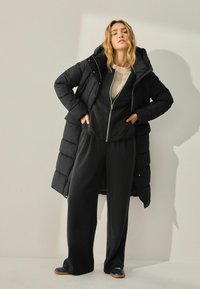 Une femme aux cheveux ondulés se tient avec assurance dans un manteau de duvet noir et une tenue sombre, les mains sur les hanches, devant un fond neutre.