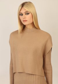 Beżowy sweter robiony na drutach z wysokim, prążkowanym kołnierzem i dopasowanymi prążkowanymi rękawami. Ciało ma luźny, warstwowy krój, co nadaje mu teksturowany wygląd.