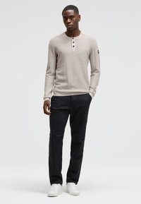 Hellbeige Langarmshirt mit Rundhalsausschnitt, dreiknöpfiger Knopfleiste und gerippten Bündchen, kombiniert mit dunklen Hosen und weißen Sneakers.