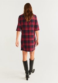 Karierter Kleid in Dunkelrot und Marineblau, kurze Ärmel, Knopfverschluss hinten, getragen mit hohen schwarzen Stiefeln, glattes Material, oberhalb des Knies.