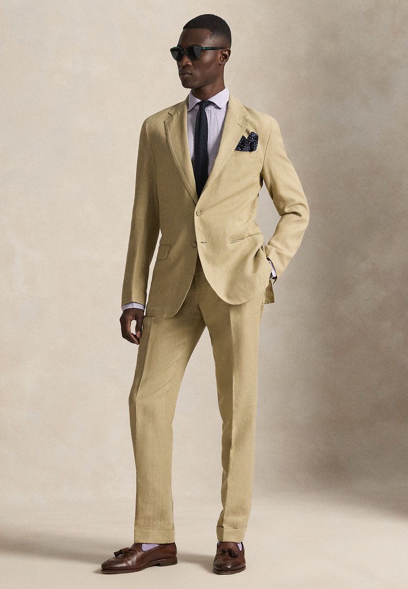 Light beige linen suit featuring a two-button jacket, notch lapels, and matching trousers. Accented with a polka dot pocket square and sunglasses.