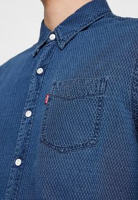 Detailní záběr na modrou texturovanou košili na knoflíky s bílými knoflíky a náprsní kapsou, na které je malá červená cedulka Levi's.