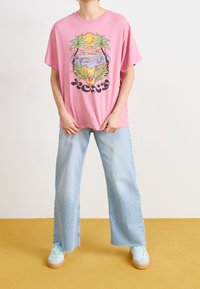 Vaaleanpunainen puuvillapaita, jossa on graafinen printti trooppisista elementeistä, yhdistettynä vaaleansinisisiin leveisiin farkkuihin ja pastellinvärisiin lenkkareihin.