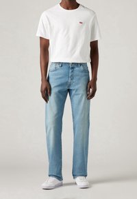 Camiseta blanca de algodón con pequeño logo rojo, combinada con jeans de mezclilla azul claro; ambos tienen un ajuste relajado y detalles mínimos. Zapatillas blancas.