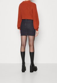 Maglione a costine rosso, mini gonna in denim scuro, collant neri trasparenti e stivali neri con tacco a blocco al ginocchio. Vista da dietro.