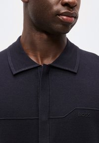 Gros plan d'une personne portant un polo côtelé bleu marine foncé avec un col et un logo "BOSS" sur la poitrine.