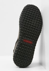 Semelle de chaussure avec motif chevron et logo rouge « Rieker » au centre, inclinée contre une surface d'angle blanche.