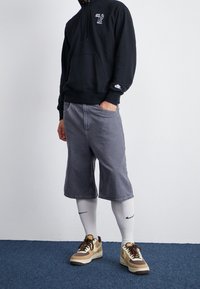 Svart huvtröja med grafik, grå knälånga shorts, vita strumpor och bruna/krämfärgade sneakers med röda detaljer.