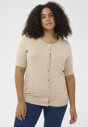 Woman with curly hair wearing a beige short-sleeve buttoned cardigan and blue jeans, standing against a plain light background.