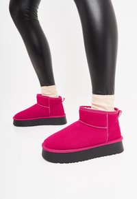 Botine roz din suede cu talpă plată neagră. Prezintă un design cu manșetă, cusături contrastante și o bandă de tragere pe călcâi.
